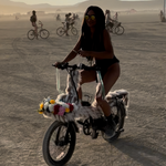 Load image into Gallery viewer, A woman rides a small electric bicycle in a desert setting at sunset, with other cyclists visible in the background. The bicycle has a white frame and handlebar wrap with colorful decorative elements.
